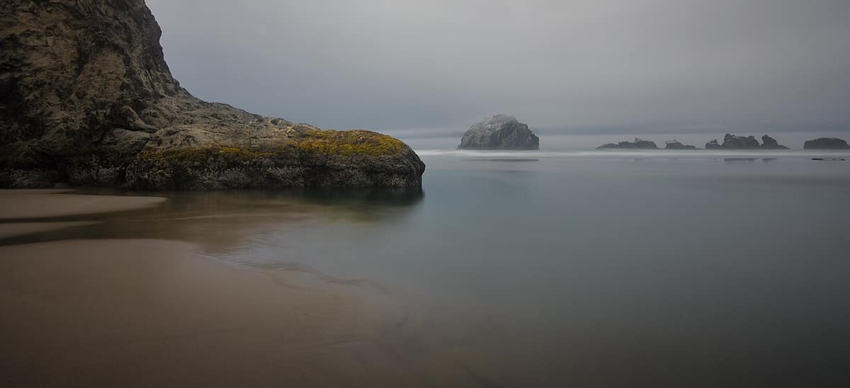A long-exposure view of a sea stack and calm tide pools on a foggy Bandon beach