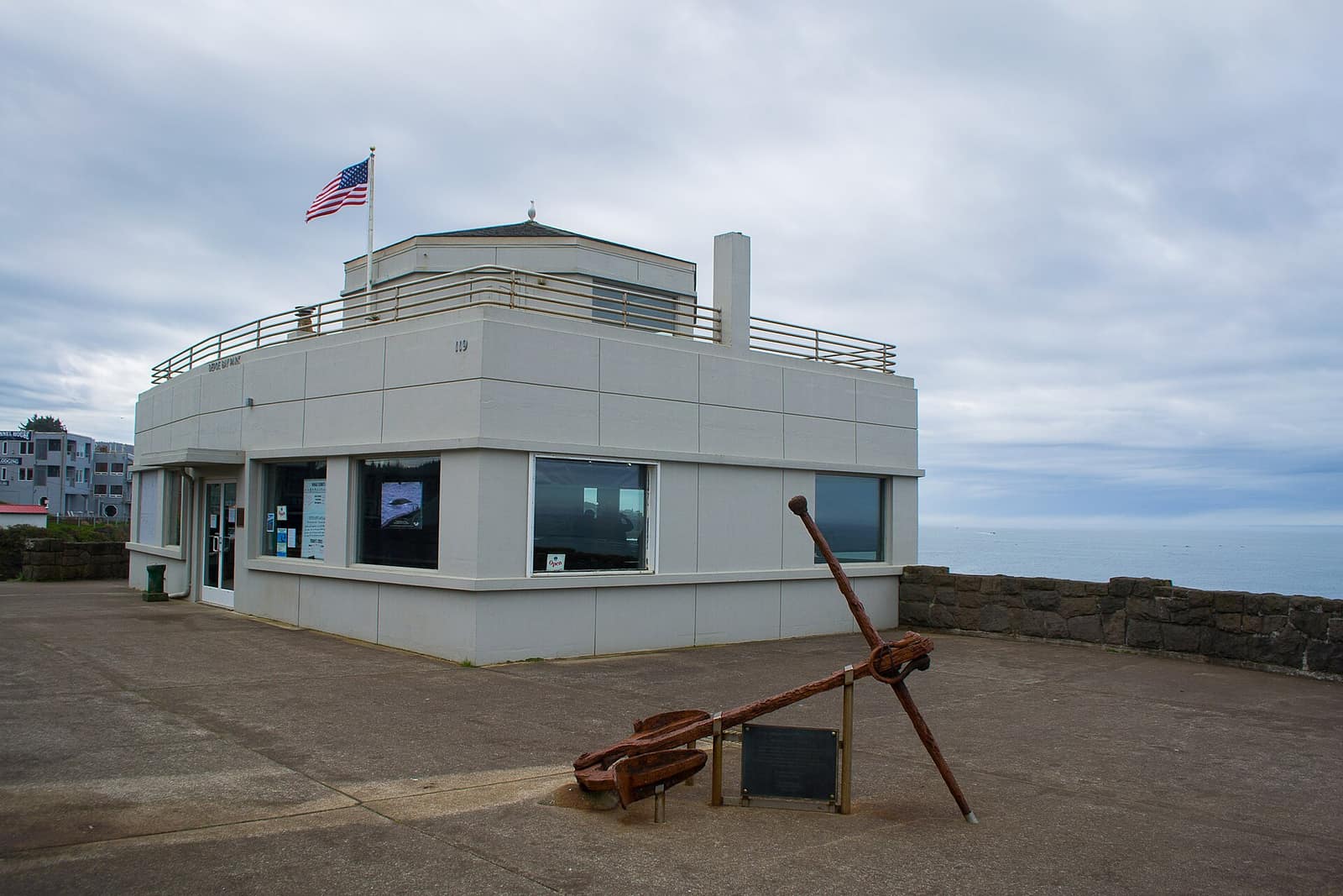 The Depoe Bay Whale Watching Center, a low gray building with a flag, and a large rusted anchor on the paved overlook in front of it