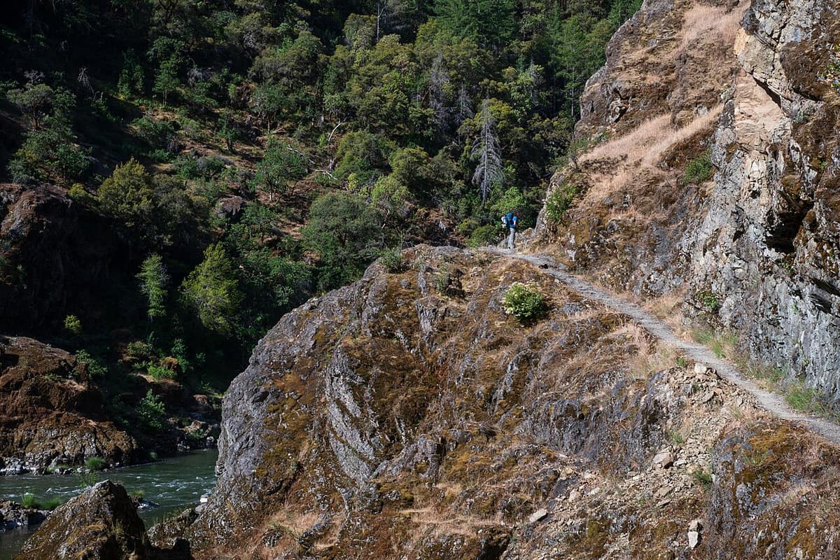 hiker on a narrow cliffside section of the Rogue River Trail above the river
