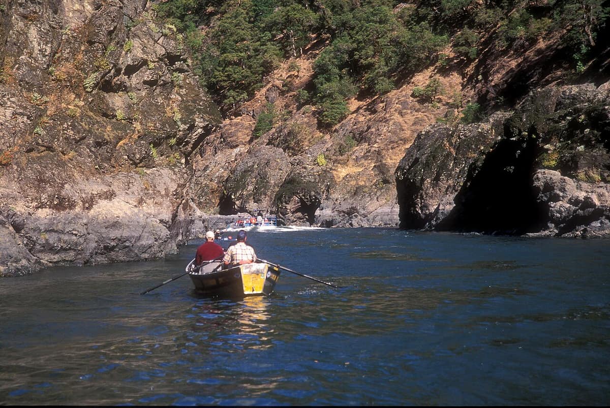 A drift boat on the Rogue River with a jet boat in the distance beneath steep rocky canyon walls