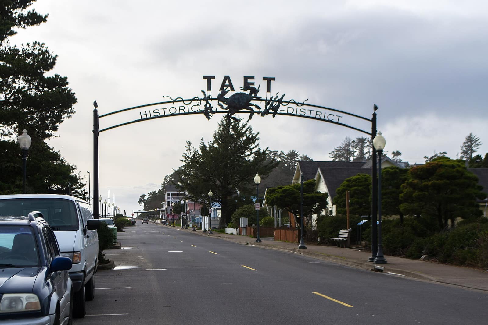 The black iron arch of the Taft Historic District over a Lincoln City street, with a crab at its center and shops and houses beyond