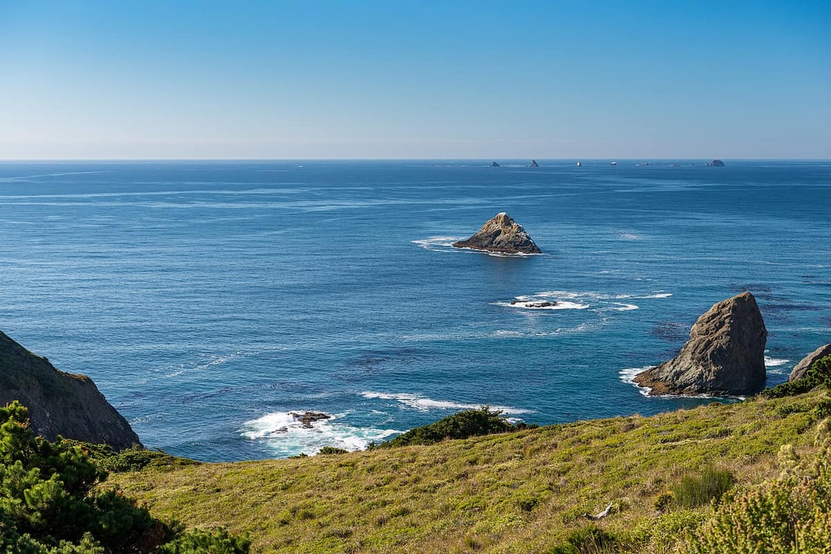 A wide sandy beach below steep green and dark rock cliffs on the coast near Port Orford, with sea stacks offshore