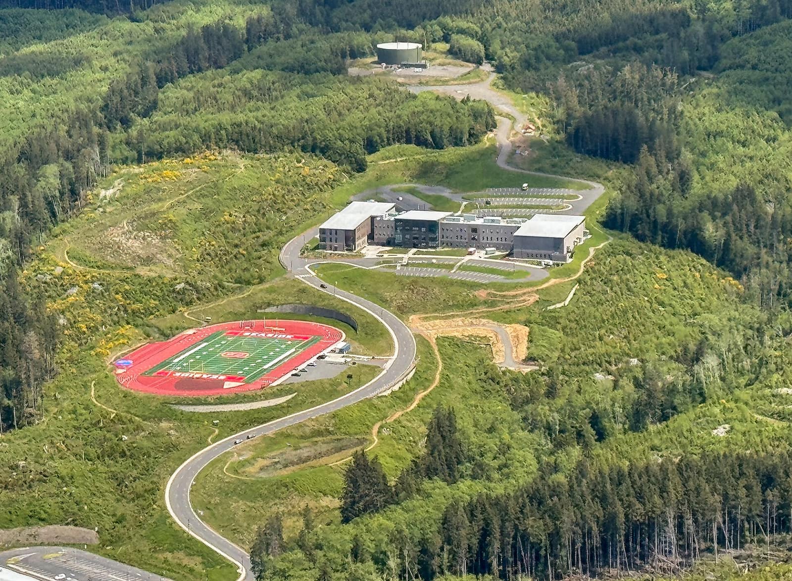 Aerial view of the hillside Seaside school campus, with a red running track and green football field below a gray school building, surrounded by forest