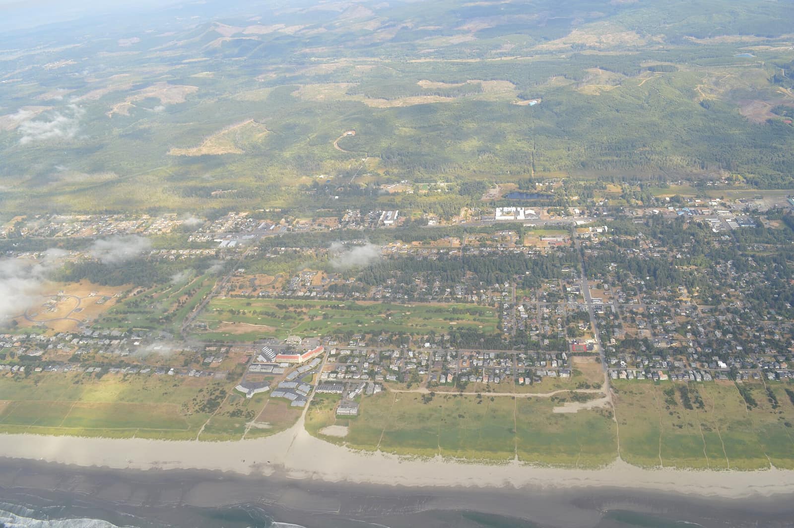 Aerial view of the town of Gearhart, with houses and a golf course behind the dunes and beach, and forested hills in the distance